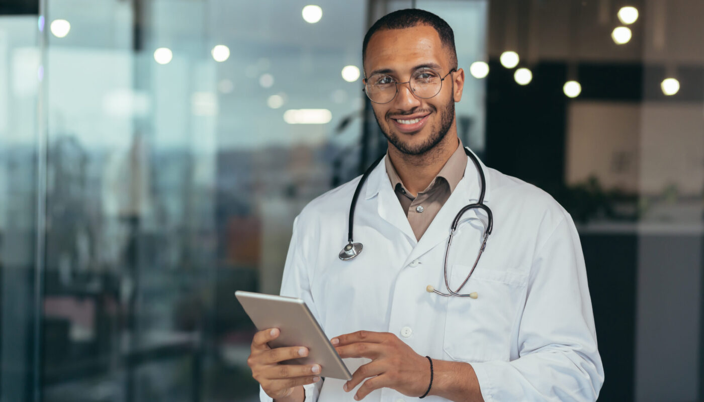 Portrait of happy and successful african american doctor man working inside office clinic holding tablet computer looking at camera and smiling wearing white coat with stethoscope.