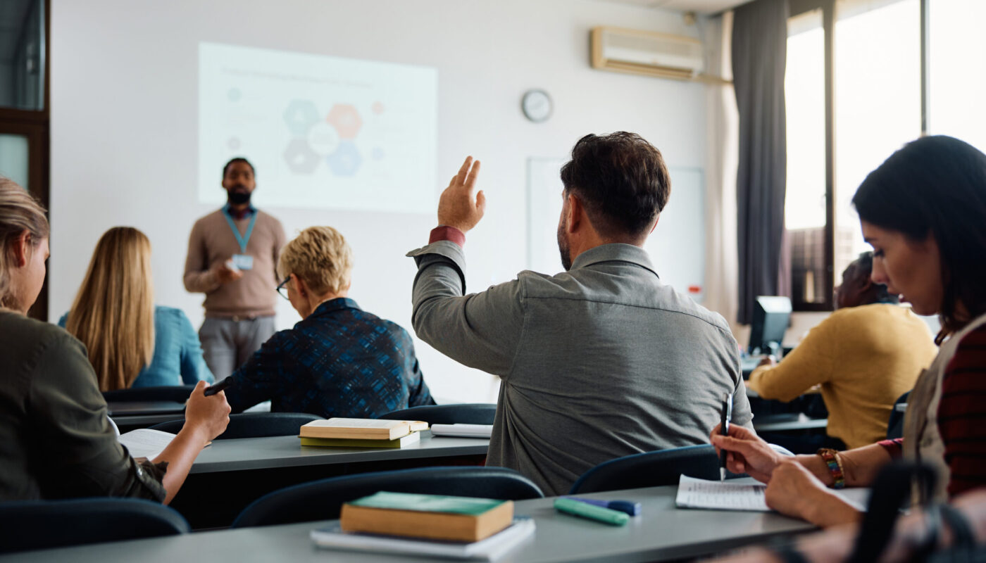 Rear view of man raising arm to ask a question during a presentation in lecture hall.